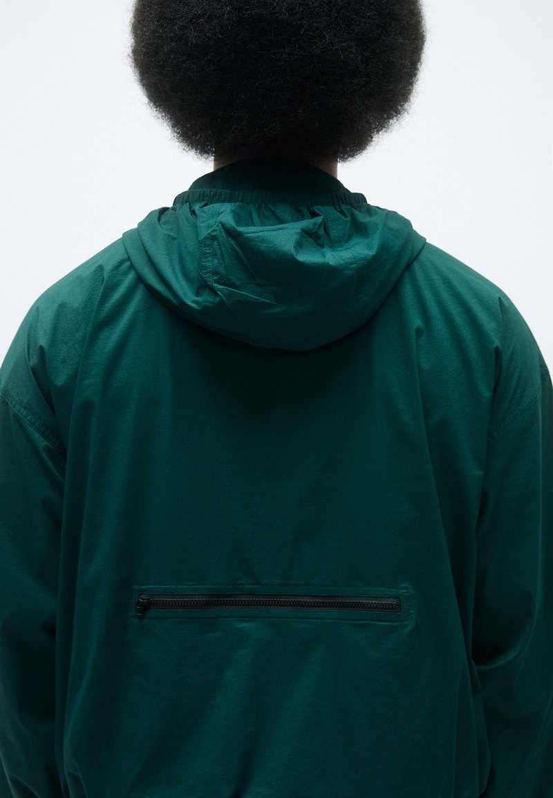 Grüne Kapuzenjacke mit glatter Textur, ausgestattet mit einer Rückenreißverschlusstasche und elastischen Bündchen. Schlichte, funktionale Gestaltung.
