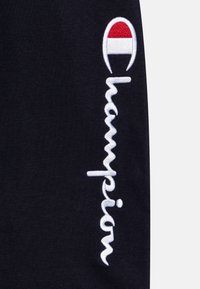 Le tissu noir présente un texte brodé en blanc écrivant "Champion" et un logo circulaire avec des accents rouges et blancs.