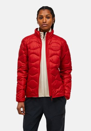 Woman wearing a red quilted jacket over a white zip-up top and black pants, standing with hands relaxed by sides against a plain background.