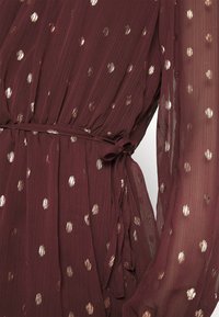 Vinigärsfärgad klänning med genomskinligt tyg, som har metalliska guldprickar, ett knytbart midjebälte och långa, flytande ärmar.