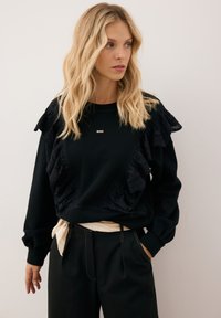 Czarna bluza z falbaniastymi akcentami i skomplikowanymi wzorami wycinanymi. Okrągły dekolt i długie rękawy z ściągaczami. Widoczny złoty detal z logo.