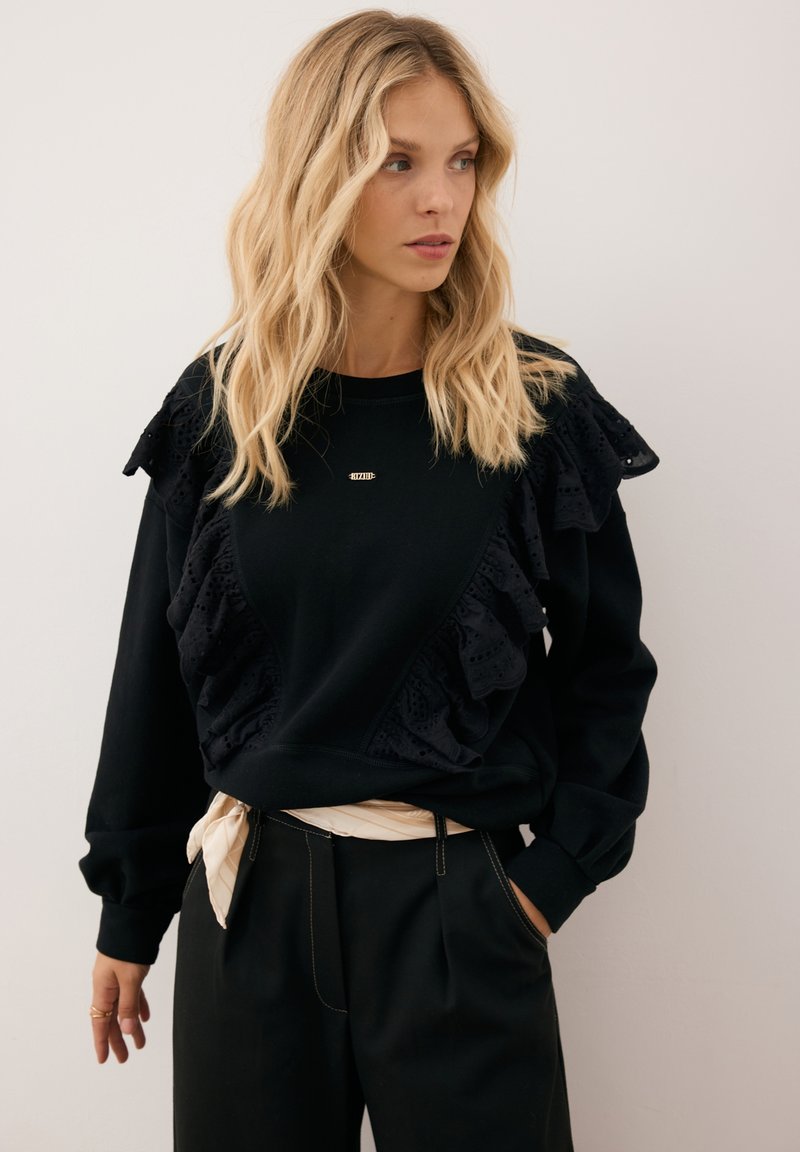 Czarna bluza z falbaniastymi akcentami i skomplikowanymi wzorami wycinanymi. Okrągły dekolt i długie rękawy z ściągaczami. Widoczny złoty detal z logo.
