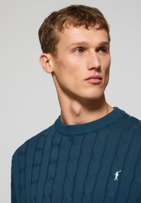 Donkerblauwe kabelgebreide trui met een ronde hals en een klein wit logo detail op de borst. Geweven patroon over de hele trui.