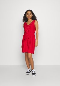 Vestido vermelho sem mangas com bordado floral na gola, cintura marcada com um laço e um corte descontraído. A modelo usa ténis pretos e está de pé sobre um fundo neutro.