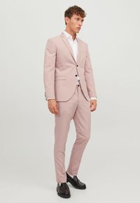 Un costume rose clair avec une coupe ajustée, une fermeture à un bouton et des revers crantés. Pantalons slim, chemise blanche en dessous et mocassins noirs.