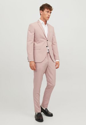 Un costume rose clair avec une coupe ajustée, une fermeture à un bouton et des revers crantés. Pantalons slim, chemise blanche en dessous et mocassins noirs.