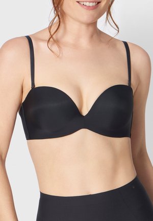 Femme souriante, portant un soutien-gorge noir sans bretelles et une culotte taille haute assortie sur un fond clair uni.