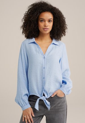 Light blue, button-up shirt with a collar, tie-front design, and long, gathered sleeves; made from textured fabric. Paired with gray jeans.