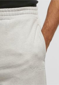 Detailný záber ruky osoby v vrecku svetlosivých teplákov s textúrovaným pásom a jemne vyšitým logom.