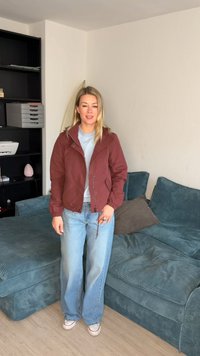 Donna bionda che indossa una giacca rossa, una camicia azzurro chiaro, jeans larghi e sneakers bianche, in piedi in un soggiorno con divano turchese e scaffalatura nera.
