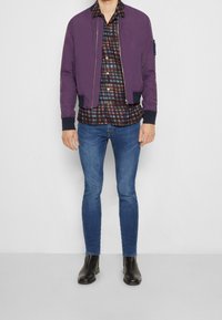 Giacca bomber viola con zip frontale, camicia a quadri con toni di blu, rosso e verde, jeans slim-fit blu e stivali neri alla caviglia.