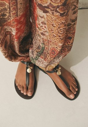 Barfüßige Füße in schwarzen Ledersandalen mit goldenen Münzanhängern, teilweise bedeckt von gemusterten, locker sitzenden Hosen auf einem beigen Boden.