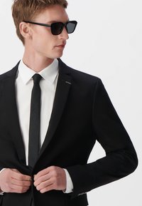 Czarny garnitur z białą koszulą i czarnym krawatem. Żakiet ma dopasowany krój, a model nosi czarne okulary przeciwsłoneczne. Gładki, strukturalny materiał.