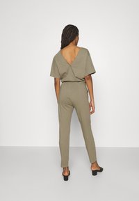 Mono verde oliva de material suave, con una espalda en V profunda, mangas cortas y piernas ajustadas. Sandalias negras planas acentúan el look.