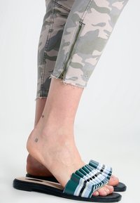 Gröna halkskydd med en veckad rem som har blå och vita ränder. Sulan är svart, och jeans med camouflage-mönster kompletterar dem.