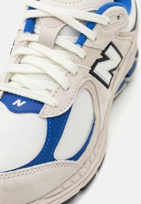 Sneaker gris clair avec des accents bleus, présentant un dessus en maille, des détails en daim, des lacets blancs et un logo brodé noir. Silhouette arrondie.