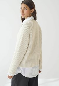 Cremefarvet strikket sweater med et struktureret mønster, dropped shoulders og ribbet kant, lagd over en hvid skjorte med sorte striber.