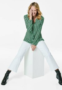 Blusa verde a maniche lunghe con un motivo geometrico, abbinata a jeans bianchi a gamba dritta e stivali neri alla caviglia, seduta su un piedistallo bianco.