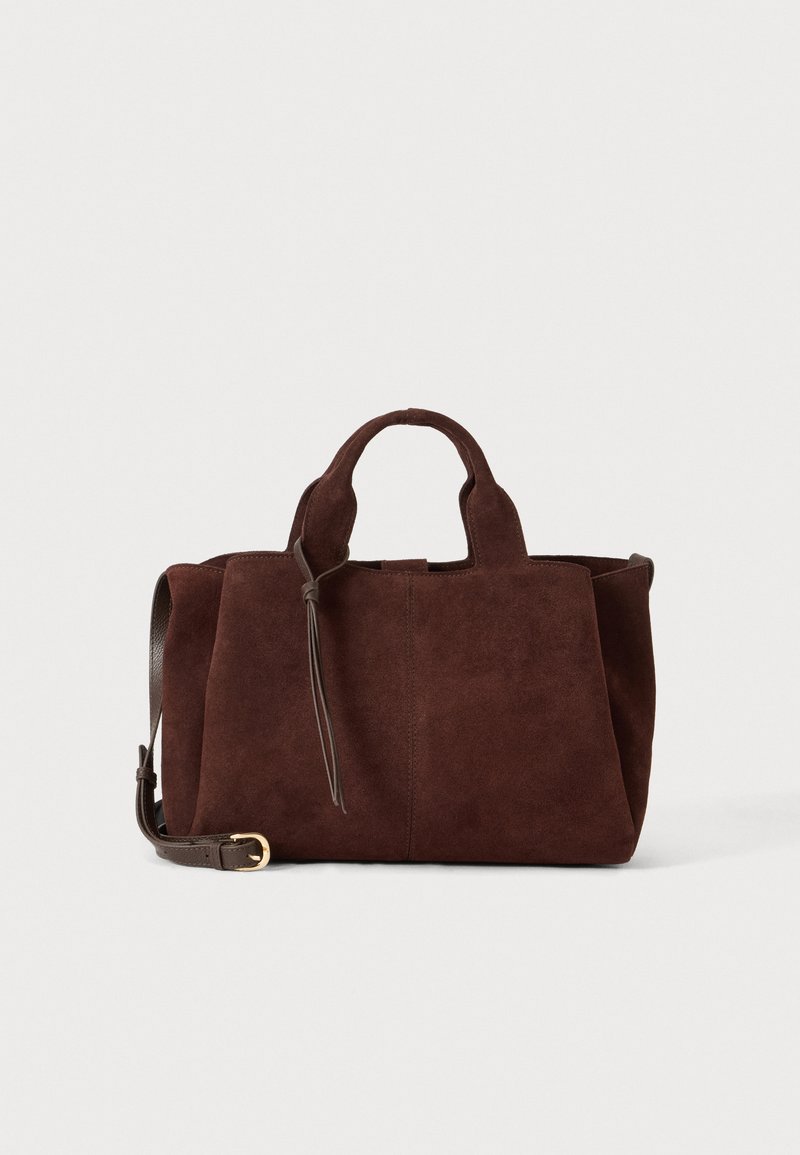 Sac à main en daim brun foncé avec des poignées courtes, une bandoulière amovible avec boucle dorée, et un simple détail pompon sur un fond uni.
