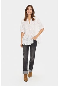 Weiße Bluse aus Lochstickerei mit V-Ausschnitt, kurzen Puffärmeln, gewelltem Saum und floralem Muster. Kombiniert mit dunklen Jeans und braunen Stiefeletten.