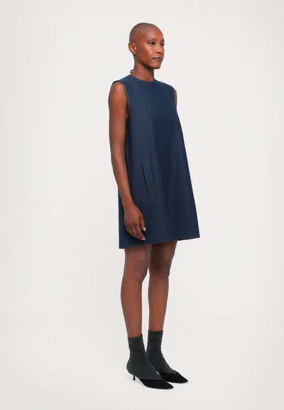 Jūras zila bez piedurknēm, brīvas formas kleita ar sānu kabatām un gludu audumu, kas apvienota ar pelēkiem zeķēm un melniem papēžiem.