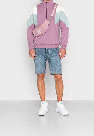 Sudadera morada con cremallera y acentos en chevron blancos y verde azulado, pantalones cortos de mezclilla y zapatillas blancas con detalles en azul claro, combinadas con una riñonera rosa.