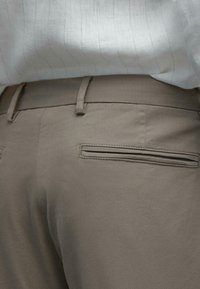 Beige katoenen broek met achterzak, ceintuurlussen, gedragen met een ingestopt lichtgekleurde shirt met dunne verticale strepen.