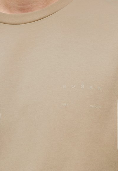 Μπεζ μπλούζα με στρογγυλή λαιμόκοψη από απαλό ύφασμα. Διαθέτει διακριτικό κείμενο: "HOGAN," "1986," και "MILANO" σε ανοιχτή απόχρωση. Ομαλή υφή.