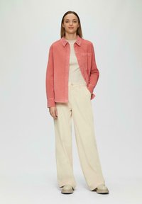 Koraal velours overhemd, witte gebreide top, beige wijde broek en crème schoenen. Het overhemd heeft een knoopsluiting aan de voorkant en een borstzak.