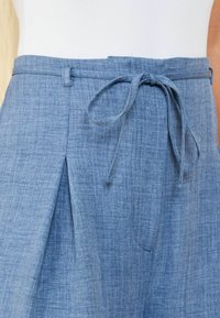 Pantalones de chambray azul con un cinturón atado, que presentan un diseño texturizado y tejido, así como un detalle plisado cerca de la cintura para un ajuste relajado.