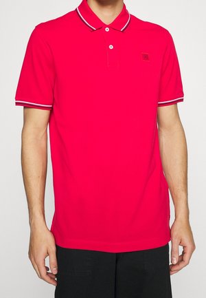 Mand iført en lyse rød kortærmet poloshirt med hvidt og sortstribet kant på krave og ærmer, kombineret med sorte bukser.