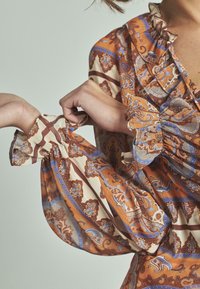 Blusa a maniche lunghe in tessuto leggero e fluido. Presenta un motivo paisley arancione, blu e crema con polsini e scollo a volant.