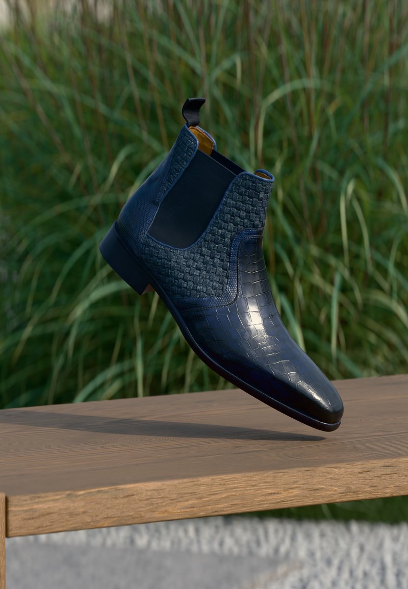 Μαύρο δερμάτινο μποτάκι αστραγάλου με υφασμένο σχέδιο και ελαστικές πλαϊνές επενδύσεις, κρεμασμένο πάνω από ένα ξύλινο παγκάκι με φόντο χορτάρι.