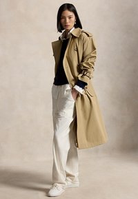 Ung kvinde iført en beige trenchcoat, hvide vidde bukser, sort sweater, hvid skjorte og hvide sneakers, stående mod en neutral baggrund.