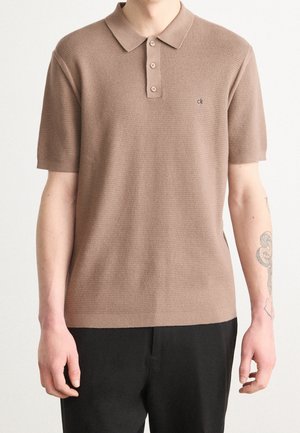 Mand iført en beige kortærmet strikket poloshirt med knapper og sorte bukser, der viser en tatovering på højre underarm.