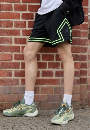 Jambes portant un short noir avec une bordure vert néon et des baskets de sport vertes avec des chaussettes blanches, debout contre un mur en brique rouge.