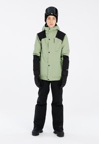 Grüne Skijacke mit schwarzen Akzenten, ausgestattet mit einer Reißverschlusstasche, kombiniert mit schwarzen Schnee-Hosen und Handschuhen, getragen über Skistiefeln und einer Skibrille.