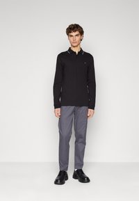 Polo a maniche lunghe nera con dettagli bianchi sul colletto, abbinata a pantaloni grigi e scarpe nere. Design semplice e aderente, con branding minimo visibile.