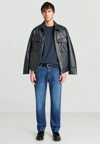 Giacca di pelle nera con due tasche sul petto, maglietta blu navy a girocollo, jeans blu a gamba dritta e scarpe nere in un ambiente neutro.