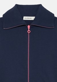 Giacca zip-up blu navy con una texture morbida, un tirazip a forma di cuore rosa e un design del colletto strutturato. Etichetta del marchio visibile all'interno.