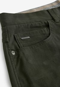 Pantalon vert texturé avec une poche avant arborant une petite étiquette métallique. La ceinture est doublée d'un tissu plus léger.