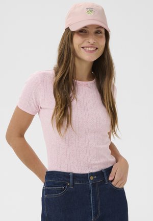 Femeie cu păr lung și castaniu, care poartă un top roz cu mâneci scurte și model, blugi albastru închis și o șapcă roz cu broderie „Matcha Girl”.