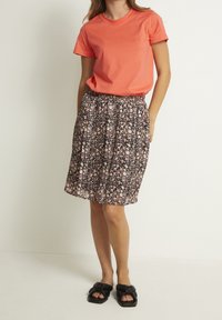 Femme portant une chemise corail à manches courtes, une jupe mi-longue à motifs floraux et des sandales plates noires avec des détails en nœud, se tenant contre un mur blanc.