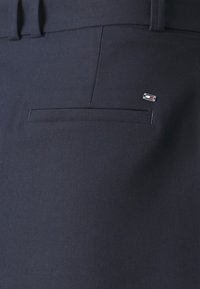 Pantaloni blu navy con una texture liscia, dotati di una tasca posteriore e un piccolo logo ricamato in rosso e bianco sul bordo della tasca.