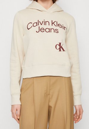 Roomkleurige hoodie met "Calvin Klein Jeans" en "cK" logo's in donkerrood, gedragen met camelkleurige, hooggetailleerde broek.