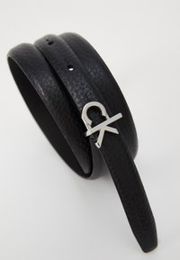 Zwarte leren riem met een gestructureerde afwerking, voorzien van een zilveren metalen gesp en het "CK"-logo. Verstelbaar met verschillende gaatjes voor maatvoering.