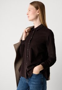 Woman with straight light brown hair wearing dark brown buttoned shirt and blue jeans, holding a beige coat over shoulder, hand in pocket.