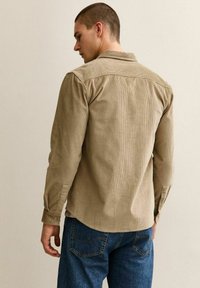 Homme aux cheveux courts portant une chemise à carreaux beige à manches longues et un jean bleu, debout face à un mur clair uni.