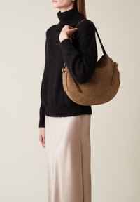 Borsa marrone testurizzata con una forma curva e dettagli in pelle, tenuta da una persona che indossa un maglione nero soffice e una gonna in satin beige.