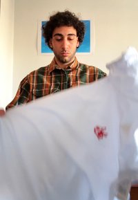 White t-shirt with a red logo, held in front of a person wearing a plaid, green and orange button-up shirt. Soft fabric, short sleeves.